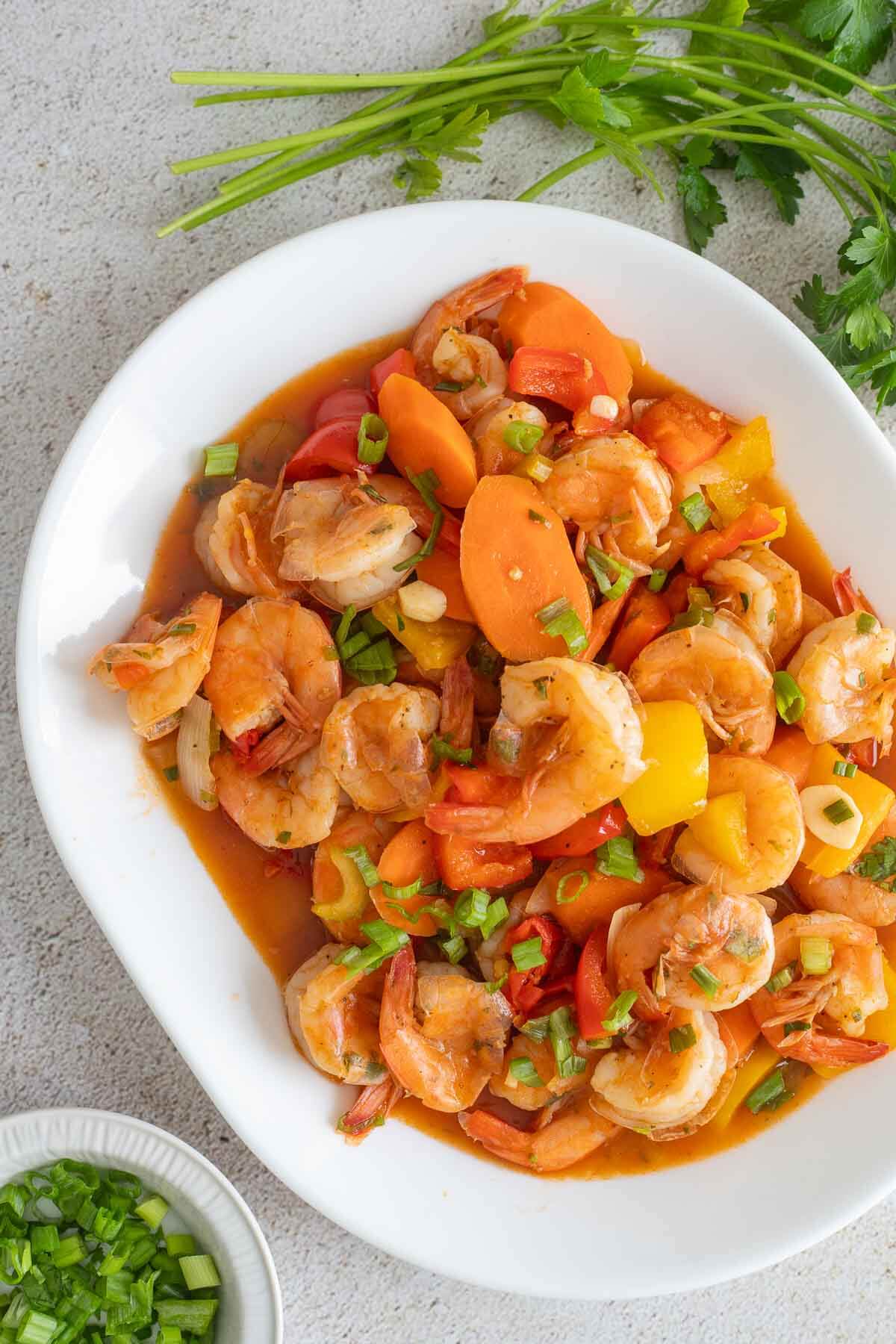 An overhead view of a white plate filled with pepper shrimp and bowl of chopped scallions on the side to garnish.