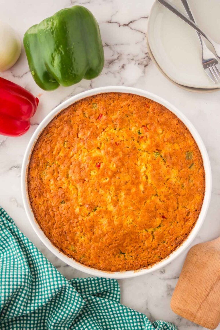 Baked trini corn casserole, in a circular white dish with a green bell peppers, plates and napkin