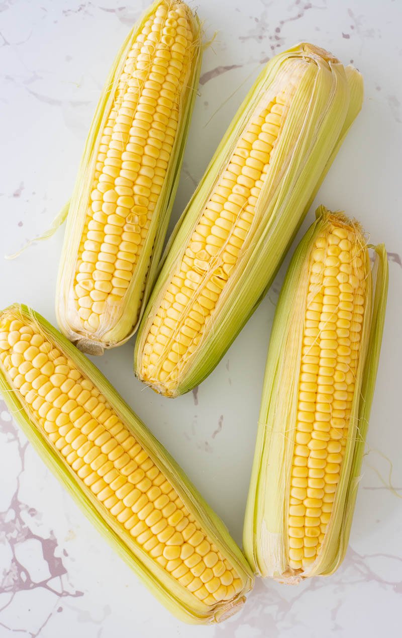 Four ears of fresh corn with silk and husks on a white counter top.