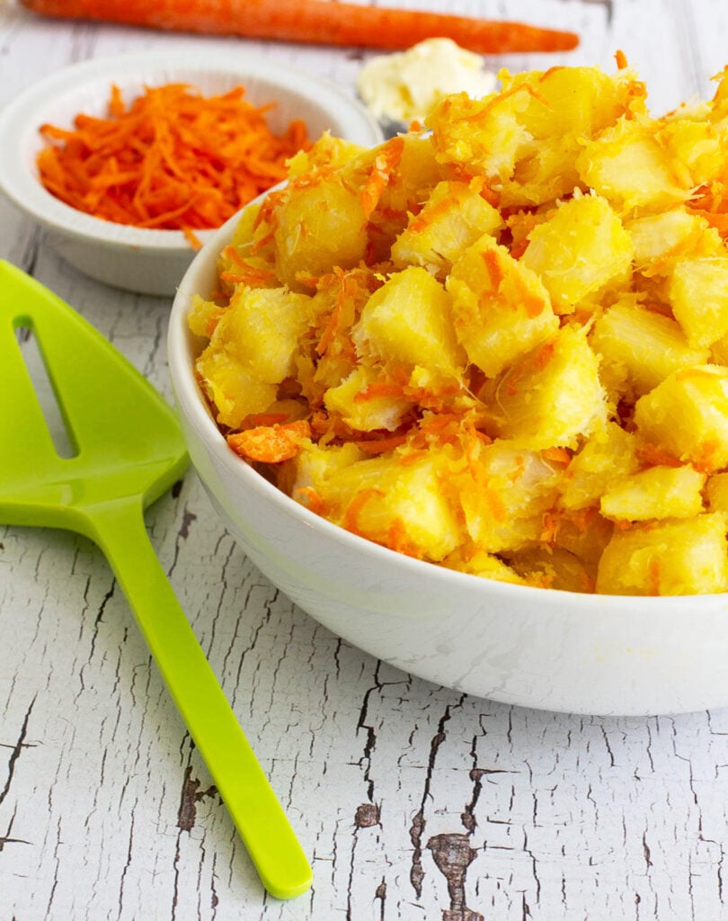 A white bowl full with butter cassava with a green spatula and a white bowl of shredded carrots in the background.