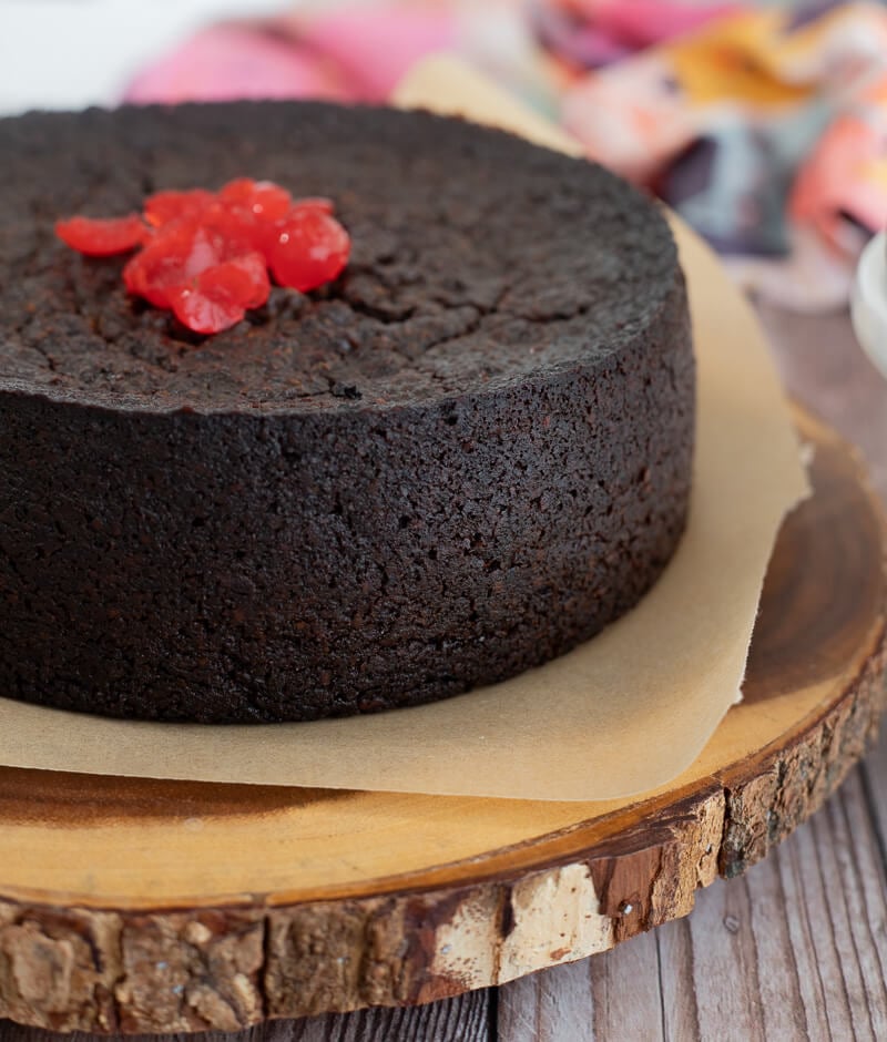 Baked dasheen black cake topped with chopped cherries on brown parchment paper.