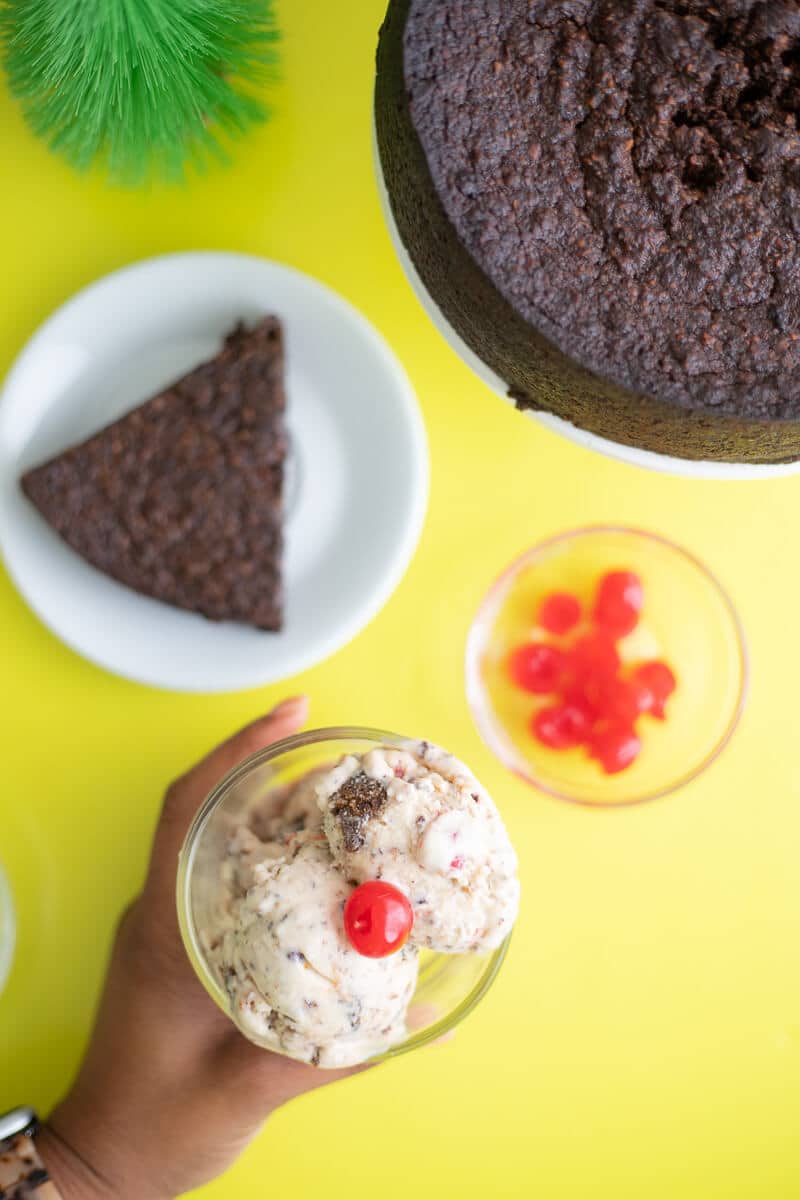 Hand holding a glass of Ice Cream with Trinidad black cake slice in background.