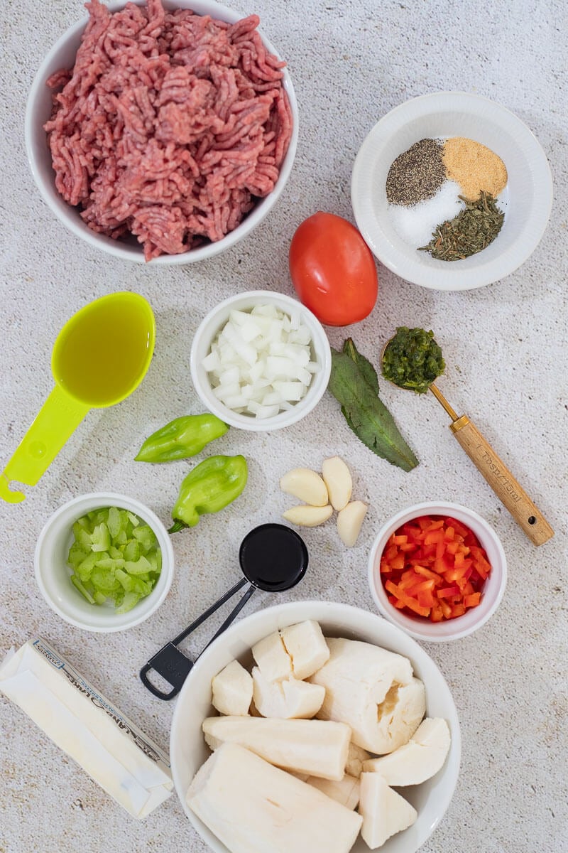Overhead view of ingredients including ground beef, cassava pieces, fresh herbs, garlic, and seasonings arranged on a white surface.