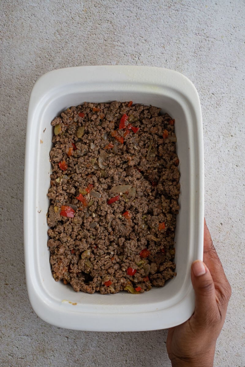 Hand holding a baking dish with the beef filling spread evenly as the base layer for cassava shepherd’s pie.
