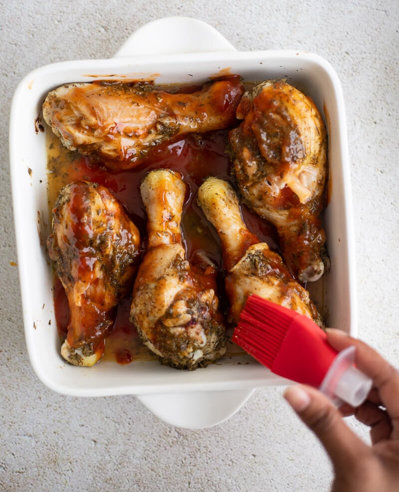 Hand using a red silicone brush to spread BBQ glaze over Trinidad baked chicken.