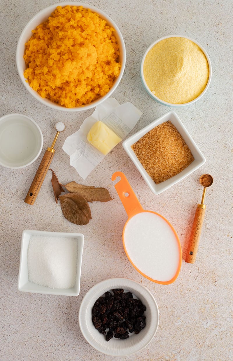 Ingredients for Trinidad cornmeal pone laid out on a light surface, including grated pumpkin, fine cornmeal, coconut milk, butter, brown sugar, and spices