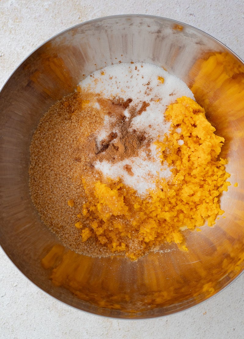 Grated pumpkin, brown sugar, white sugar, and Caribbean spices combined in a mixing bowl to start the cornmeal pone batter.