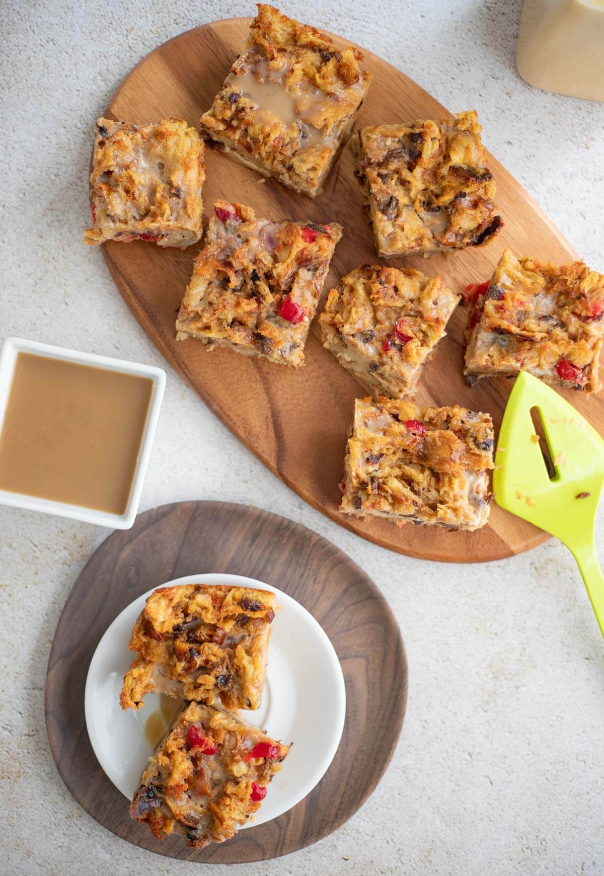 Overhead view of sliced Ponche de Crème Bread Pudding on a wooden tray with glaze in a bowl and two slices served on a white plate.
