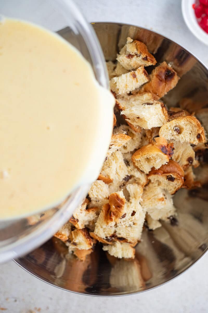 Pouring custard over cubed challah bread in a metal mixing bowl.