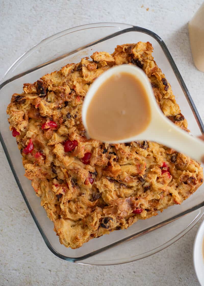 Ladle pouring creamy ponche de crème glaze over freshly baked pudding with cherries and raisins in a glass baking dish.
