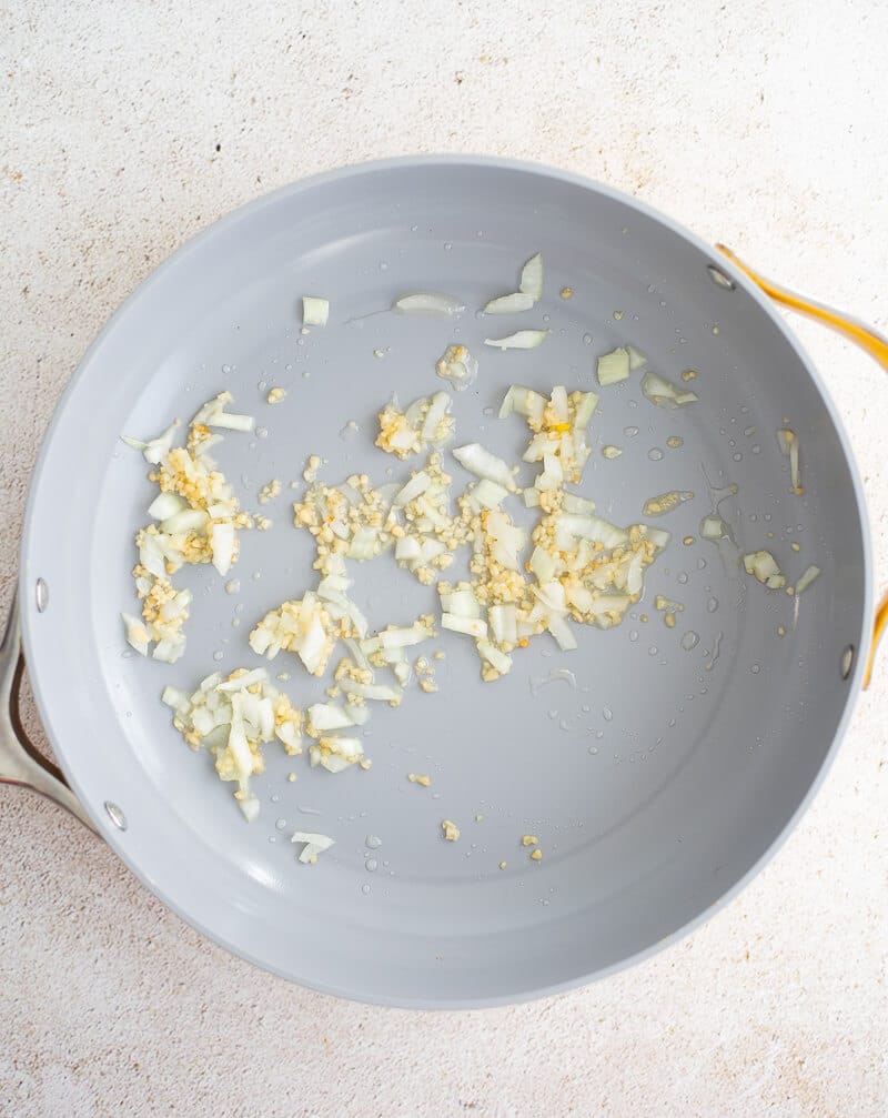 Garlic and chopped onion sautéing in oil in a gray skillet.