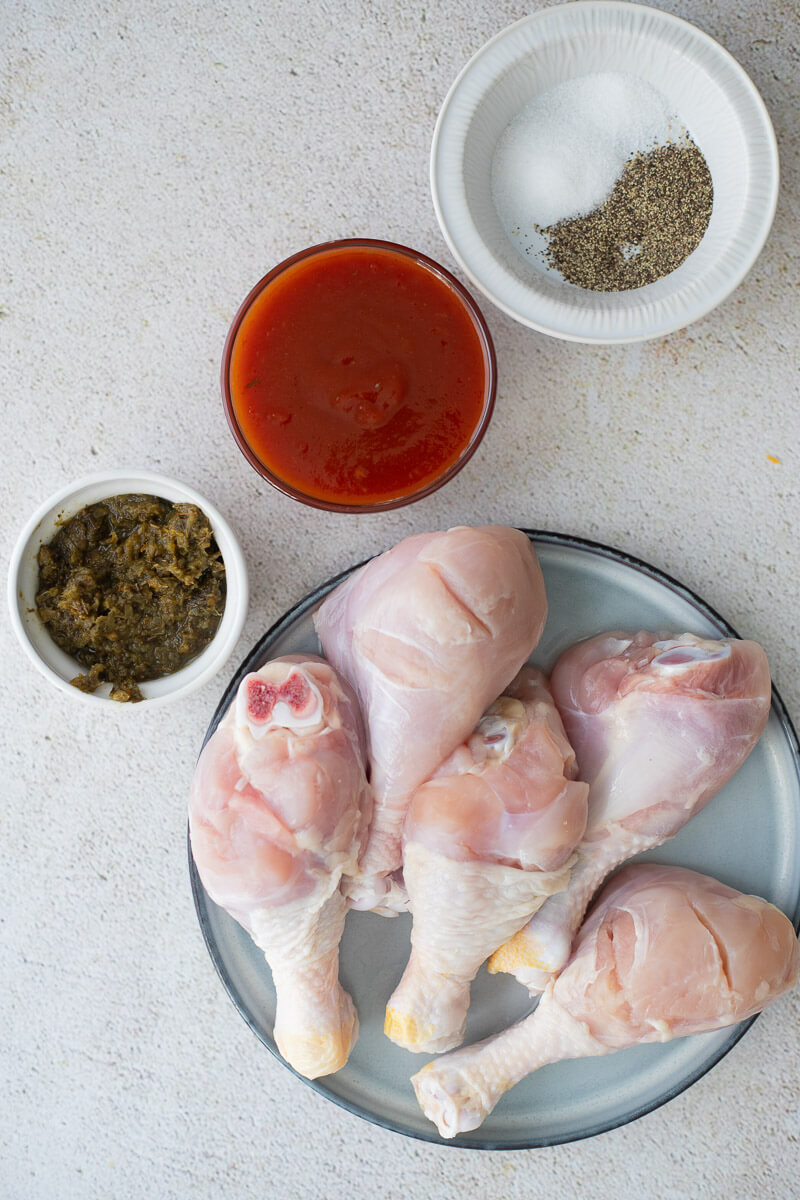 Ingredients for Trinidad-style baked chicken including raw drumsticks, green seasoning, BBQ sauce, and salt and black pepper.