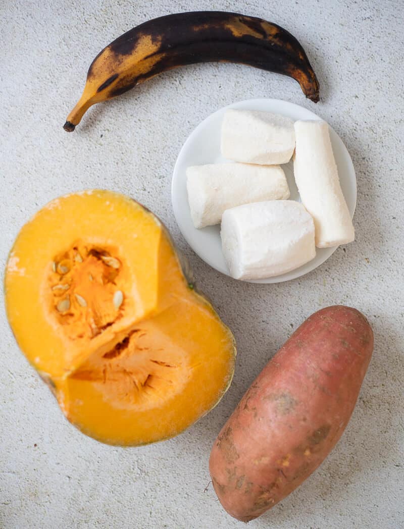 Caribbean ground provisions including ripe plantain, frozen cassava pieces, pumpkin, and sweet potato arranged on a light background.