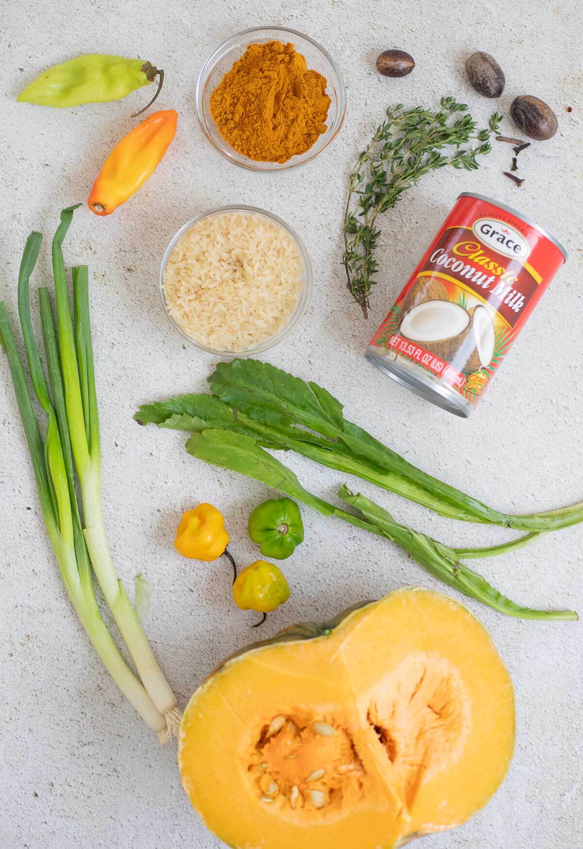 Flat lay of Caribbean pantry staples including coconut milk, curry powder, rice, hot peppers, thyme, nutmeg, chadon beni, scallions, and pumpkin.