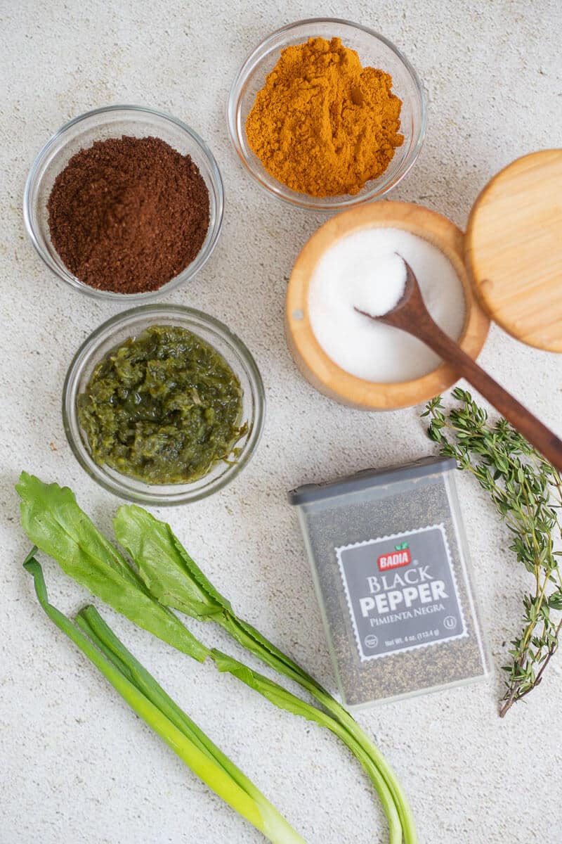 Caribbean cooking essentials including curry powder, ground geera, green seasoning, black pepper, salt, thyme, chadon beni, and scallions on a kitchen counter.