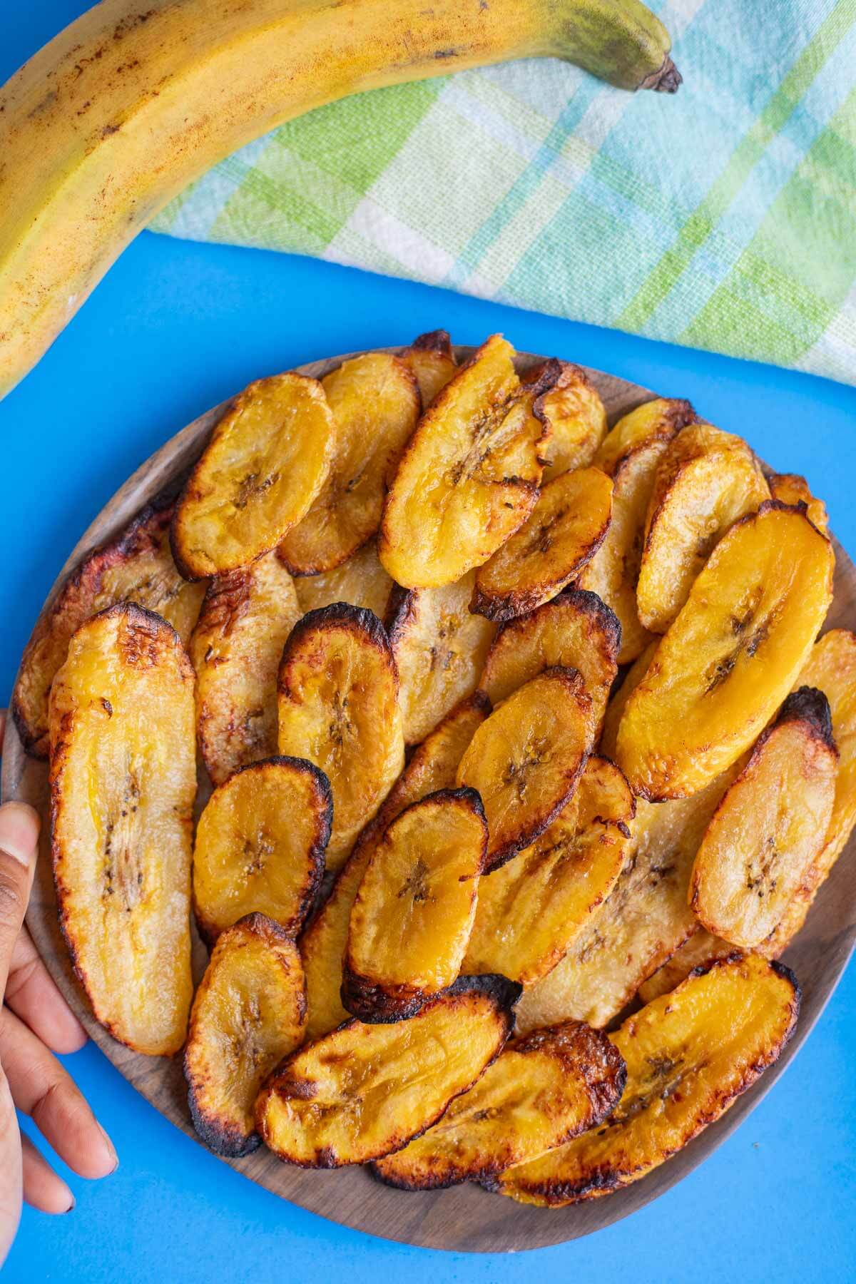 Overhead shot of a plate of baked plantains with a ripe plantain and green checked napkin on a blue surface.