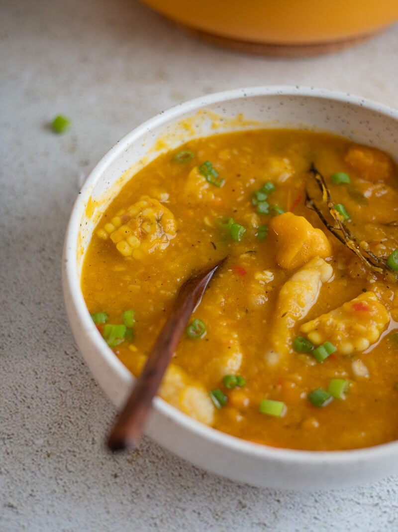 A close-up of hearty Jamaican pumpkin soup with dumplings and pumpkin pieces, served with a wooden spoon.