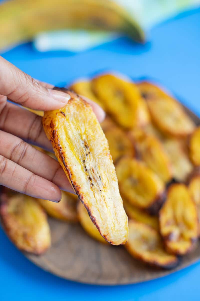 Close-up of a hand holding a slice of golden baked plantains with more slices blurred in the background.