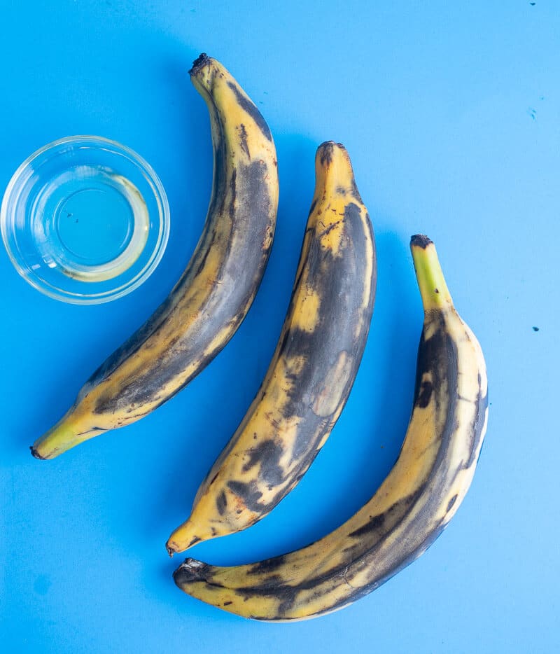 Three ripe plantains with black spots beside a small bowl of oil on a bright blue background.