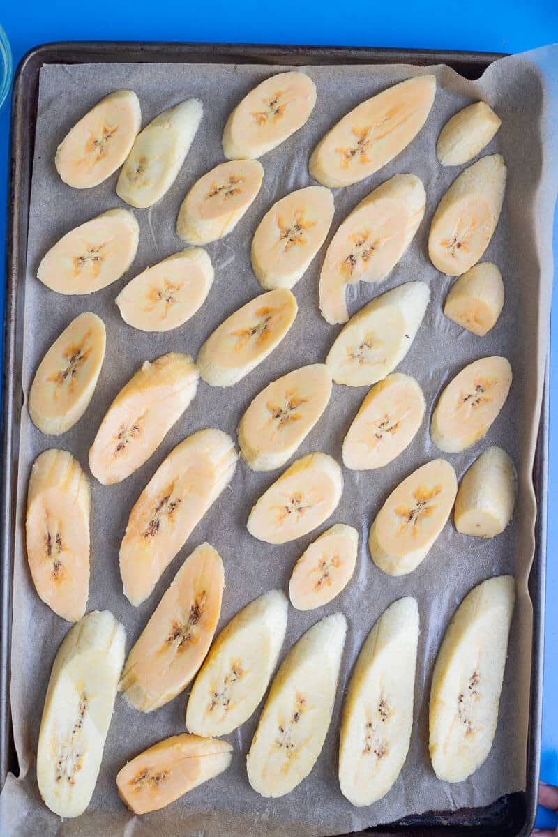 Overhead of peeled, sliced ripe plantains arranged on a parchment-lined baking tray before baking.