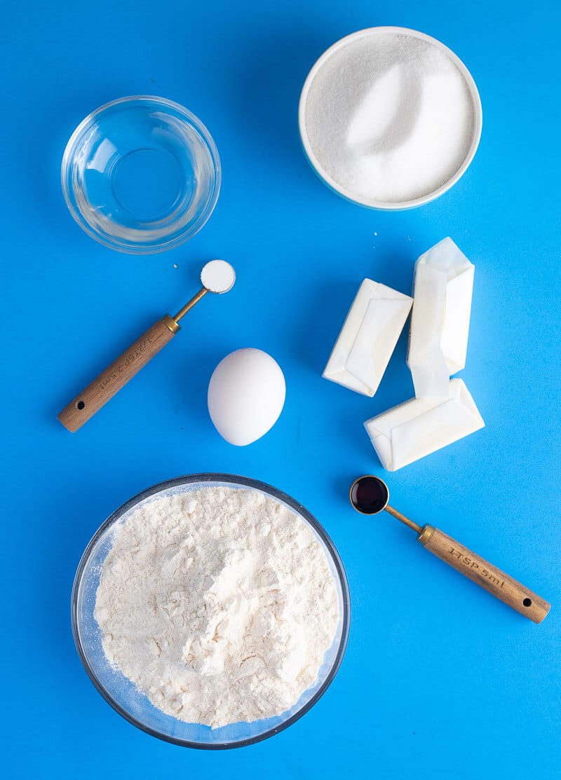 Ingredients for cookies including cassava flour, sugar, margarine, egg, vanilla, baking powder, and water on a blue background