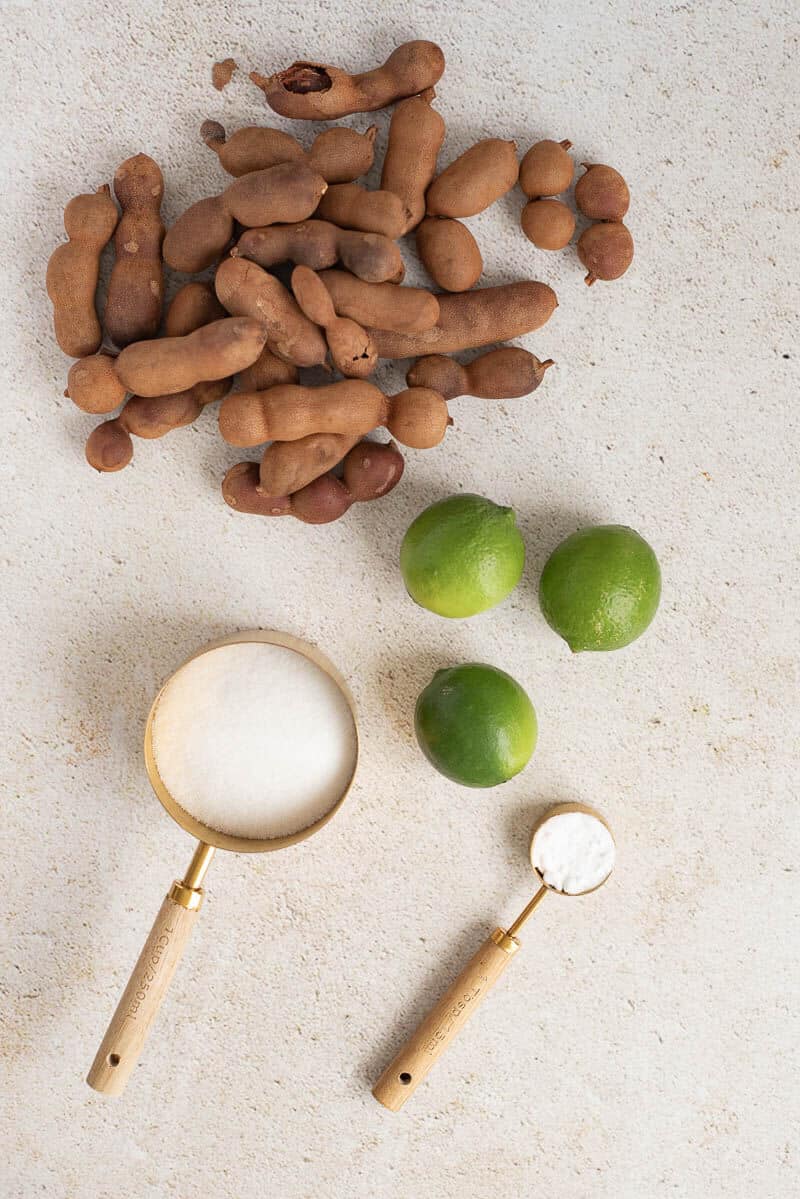 Ingredients for homemade tamarind juice including fresh tamarind pods, sugar, baking soda, and limes.