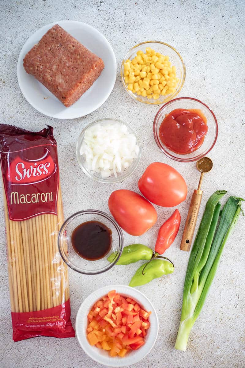 Ingredients for macaroni and corned beef including canned corned beef, pasta, vegetables, ketchup, and seasonings